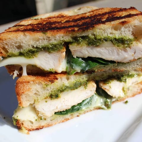 Ein saftiger Chicken Pesto Panini liegt auf einem hölzernen Schneidebrett, gefüllt mit Tomaten und Rucola und einer leicht geöffneten Hälfte.
